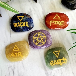 May include: Five polished stones with gold lettering and symbols representing the elements: Water, Fire, Air, Earth, and a pentagram. The stones are blue, red, yellow, purple, and green. The background is a white marble surface with green plants.