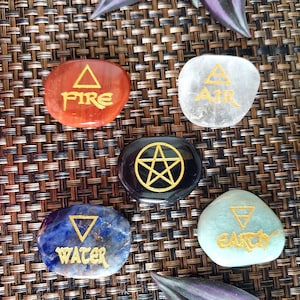 May include: A collection of polished stones, each inscribed with a symbol and a word representing the elements: Fire, Air, Water, and Earth. The stones are in various colors, including red, clear, blue, and green, with gold lettering. A black stone features a gold pentagram.