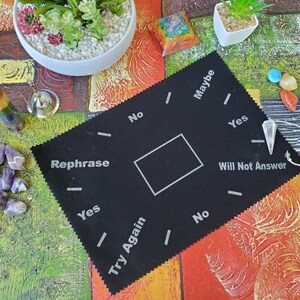 May include: Black felt pendulum board with the words "Yes", "No", "Maybe", "Rephrase", "Try Again", and "Will Not Answer" printed on it. A white pendulum is hanging from a string.