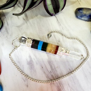 May include: A silver chain dangles from a clear quartz crystal pendulum with a multi-colored stone base. The base features black, blue, green, yellow, red, and pink stripes.