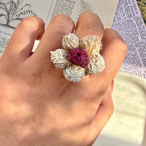 Può includere: Un anello unico con un design floreale. I petali dell'anello sono fatti di pietre strutturate beige e bianche, che circondano una pietra centrale rosa intenso. L'anello è su una mano, con carta vintage sullo sfondo.