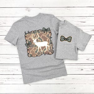 May include: Two gray t-shirts, one adult and one child size, with matching deer-themed designs. The adult shirt features a white deer silhouette on a camouflage background, framed with bows. The child's shirt has a bow tie.