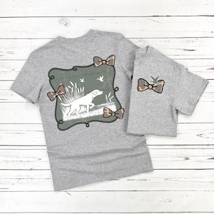 May include: Two gray t-shirts, one adult and one child size, with matching hunting-themed designs. The adult shirt features a large graphic of a dog in a field, framed by decorative bows. The child's shirt has a smaller bow design.