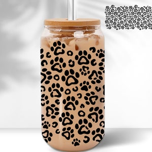 May include: A clear glass reusable water bottle with a bamboo lid and a black paw print pattern. The bottle is filled with a light brown liquid.