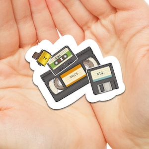 May include: A collection of vintage media storage devices, including a cassette tape, a VHS tape, and a floppy disk. The devices are arranged in a sticker format and are labeled "old."