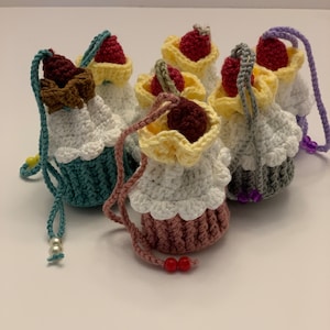May include: A collection of handmade crochet cupcake ornaments. Each cupcake has a unique color scheme, with white frosting, a colorful base, and a cherry or chocolate topping. They have a loop for hanging and are adorned with beads.
