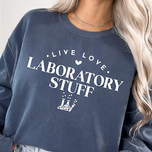 May include: A dark blue sweatshirt with the text "...LIVE LOVE. LABORATORY STUFF" in white lettering. A small heart symbol is between the words "LIVE" and "LOVE". A graphic of three beakers with bubbles is below the text.