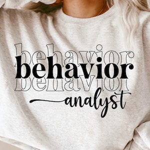 May include: A gray sweatshirt with the text "behavior analyst" printed in black. The text is layered and stacked on top of itself.
