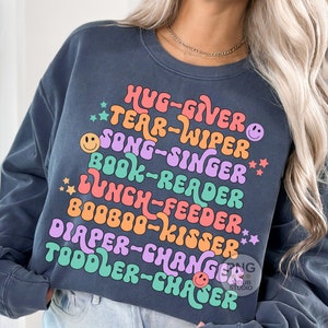 May include: A dark blue sweatshirt with a colorful graphic design that reads "Hug-Giver, Tear-Wiper, Song-Singer, Book-Reader, Lunch-Feeder, Booboo-Kisser, Diaper-Changer, Toddler-Chaser".