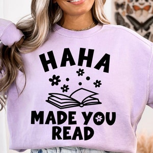 May include: A lavender sweatshirt with the text "HAHA MADE YOU READ" in black. The text is surrounded by small black flowers and a black outline of an open book.