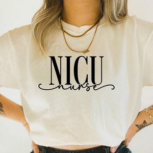 May include: Cream-colored t-shirt with the words "NICU nurse" in black, stylized font. The shirt has a crew neck and short sleeves. A gold necklace is visible at the neckline. The overall style is casual and comfortable.