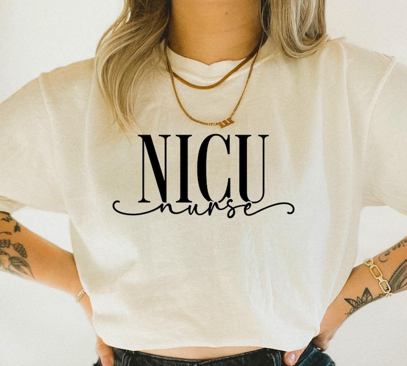 May include: Cream-colored t-shirt with the words "NICU nurse" in black, stylized font. The shirt has a crew neck and short sleeves. A gold necklace is visible at the neckline. The overall style is casual and comfortable.