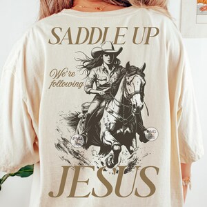 May include: Cream-coloured t-shirt with a sepia-toned graphic of a cowgirl on horseback. The text "SADDLE UP" is above the graphic, with "We're following" and "JESUS" below. The shirt has a loose fit.