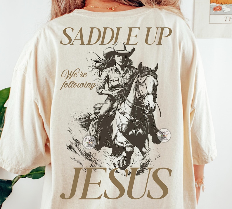 May include: Cream-coloured t-shirt with a sepia-toned graphic of a cowgirl on horseback. The text "SADDLE UP" is above the graphic, with "We're following" and "JESUS" below. The shirt has a loose fit.