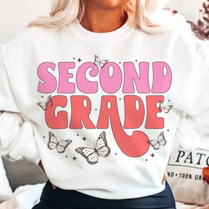 May include: White sweatshirt with the text "Second Grade" in pink and red retro font. The text is surrounded by small butterfly graphics.