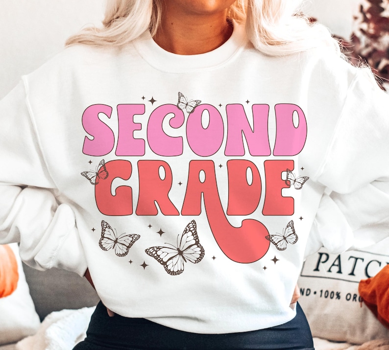 May include: White sweatshirt with the text "Second Grade" in pink and red retro font. The text is surrounded by small butterfly graphics.