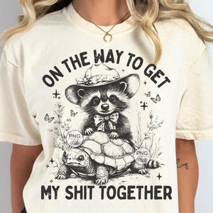 K&ouml;nnte beinhalten: Ein cremefarbenes T-Shirt mit einer schwarz-wei&szlig;en Grafik eines Waschb&auml;ren, der einen Hut tr&auml;gt und auf einer Schildkr&ouml;te reitet. Der Text "On the way to get my shit together" ist auf das Shirt gedruckt.