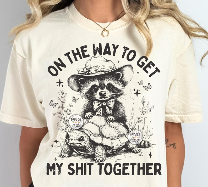K&ouml;nnte beinhalten: Ein cremefarbenes T-Shirt mit einer schwarz-wei&szlig;en Grafik eines Waschb&auml;ren, der einen Hut tr&auml;gt und auf einer Schildkr&ouml;te reitet. Der Text "On the way to get my shit together" ist auf das Shirt gedruckt.