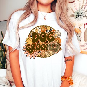 May include: White t-shirt with a brown and yellow graphic design that says "Dog Groomer" with flowers and smiley faces. The graphic is in a retro style.