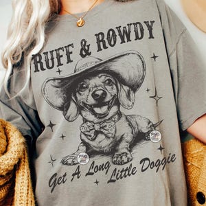 May include: A gray t-shirt with a black and white illustration of a dachshund wearing a cowboy hat. The text "Ruff & Rowdy" is above the dog and "Get A Long Little Doggie" is below the dog.