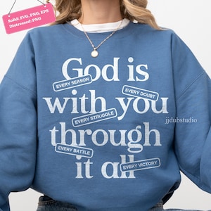 May include: A blue sweatshirt with the words "God is with you through it all" in white lettering. The text is surrounded by smaller phrases like "Every Season" and "Every Doubt". The sweatshirt has a crew neck.