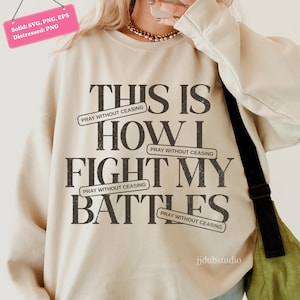 以下が含まれることがあります： 「THIS IS HOW I FIGHT MY BATTLES」の黒い文字がプリントされたクリーム色のスウェットシャツ。「PRAY WITHOUT CEASING」のバナー付き。クルーネックと長袖。画像には「jjdubstudio」の文字も含まれています。