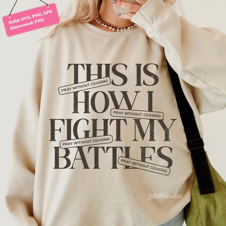 以下が含まれることがあります： 「THIS IS HOW I FIGHT MY BATTLES」の黒い文字がプリントされたクリーム色のスウェットシャツ。「PRAY WITHOUT CEASING」のバナー付き。クルーネックと長袖。画像には「jjdubstudio」の文字も含まれています。