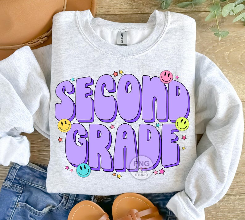 May include: A light grey sweatshirt with the text "Second Grade" in purple with a 3D effect. The text is surrounded by colorful stars and smiley faces.