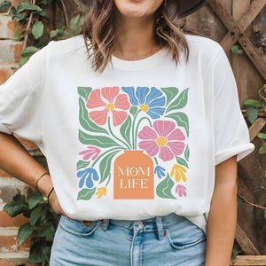 May include: White t-shirt with a floral design and the text "MOM LIFE" printed on the front. The flowers are pink, blue, and orange.