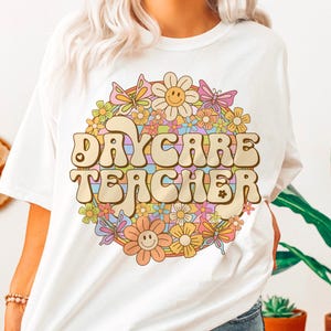 Peut inclure: T-shirt blanc avec un graphique r&eacute;tro d'un arc-en-ciel et de fleurs avec le texte "Daycare Teacher".