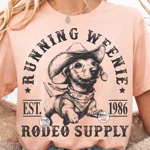 May include: A peach-colored t-shirt with a black and white illustration of a dachshund wearing a cowboy hat. The text "Running Weenie Est. 1986 Rodeo Supply" is printed on the shirt.