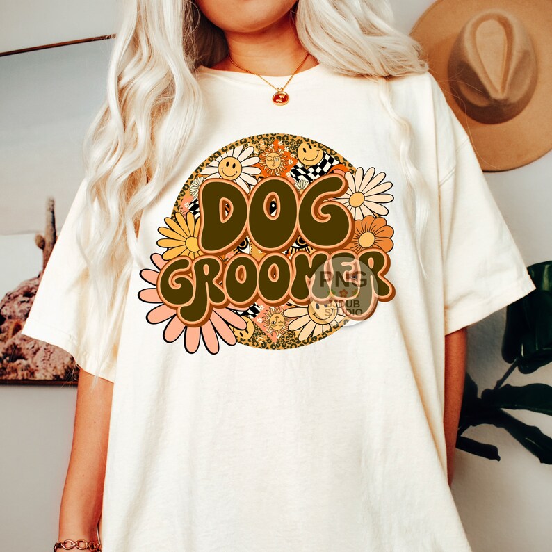 May include: A white t-shirt with a brown and orange graphic design that says "DOG GROOMER" in a retro font. The graphic is surrounded by flowers and a leopard print pattern.