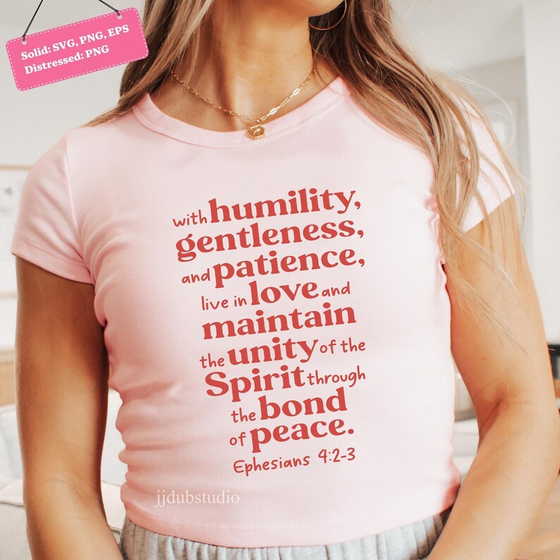 以下が含まれることがあります： 丸首と半袖のライトピンクのTシャツ。エペソ人への手紙4:2-3からの引用が赤い文字で書かれており、謙虚さ、優しさ、忍耐、愛、平和を促進しています。テキストは様式化されたフォントです。