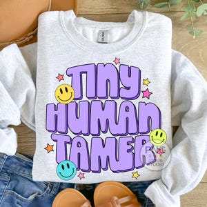 Peut inclure: Un sweat-shirt gris clair avec le texte "Tiny Human Tamer" en violet avec un smiley jaune, bleu et vert. Le texte est dans un style de police r&eacute;tro avec des &eacute;toiles roses, jaunes et orange.