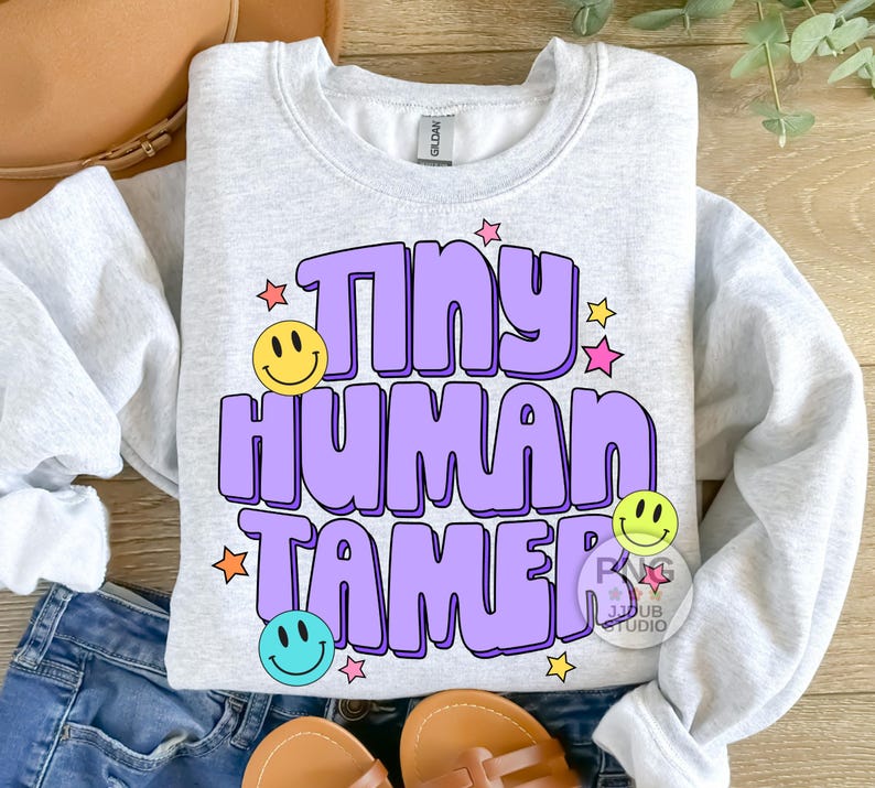 Peut inclure: Un sweat-shirt gris clair avec le texte "Tiny Human Tamer" en violet avec un smiley jaune, bleu et vert. Le texte est dans un style de police r&eacute;tro avec des &eacute;toiles roses, jaunes et orange.