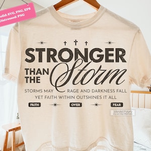 Pode incluir: Camiseta bege com a frase "STRONGER THAN THE STORM" em letras marrons escuras. O design inclui as frases "STORMS MAY RAGE AND DARKNESS FALL" e "YET FAITH WITHIN OUTSHINES IT ALL".