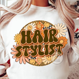 May include: White sweatshirt with a brown and orange retro-style graphic that says "Hair Stylist" surrounded by daisies and smiley faces.