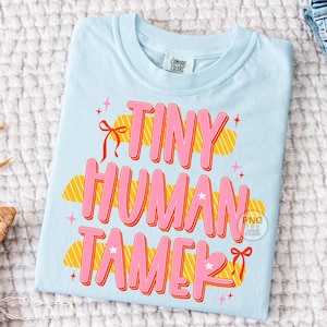 以下が含まれることがあります： ピンク色の「TINY HUMAN TAMER」の文字がオレンジ色で縁取られた、水色のTシャツ。デザインには星、リボン、黄色とオレンジ色の雲のような形が含まれています。Tシャツは白いテクスチャのある表面に折りたたまれています。