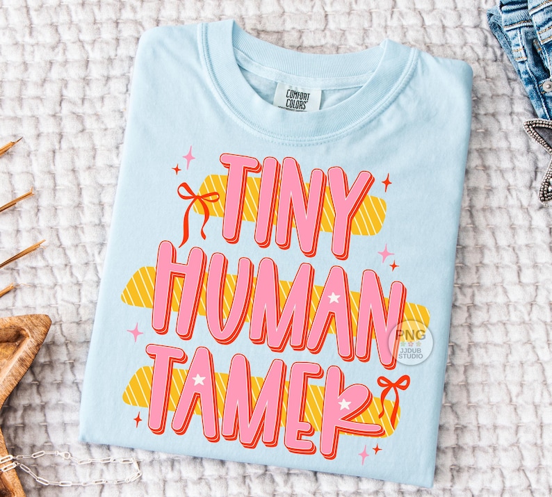 以下が含まれることがあります： ピンク色の「TINY HUMAN TAMER」の文字がオレンジ色で縁取られた、水色のTシャツ。デザインには星、リボン、黄色とオレンジ色の雲のような形が含まれています。Tシャツは白いテクスチャのある表面に折りたたまれています。