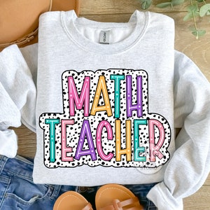 Op de afbeelding: Een grijs sweatshirt met de tekst "MATH TEACHER" in kleurrijke letters op een zwart-witte stippenachtergrond.