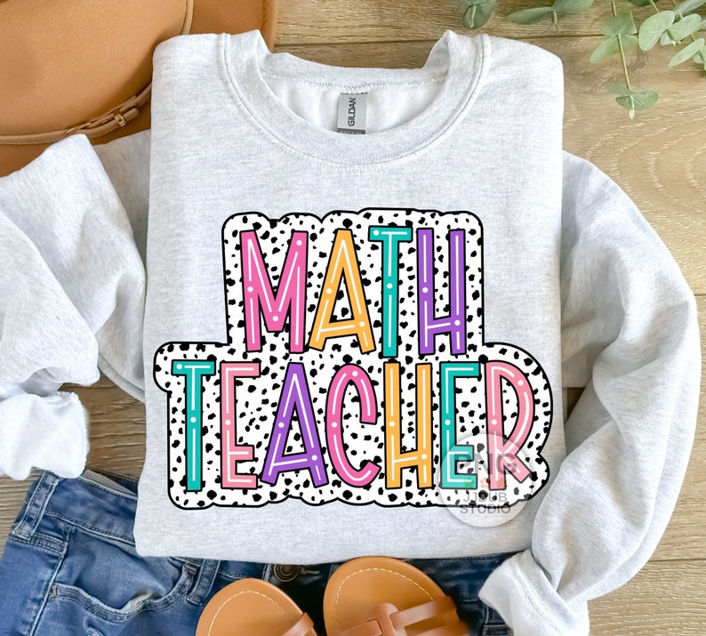 Op de afbeelding: Een grijs sweatshirt met de tekst "MATH TEACHER" in kleurrijke letters op een zwart-witte stippenachtergrond.