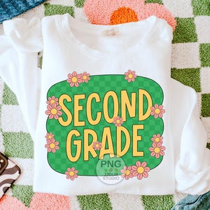 May include: White sweatshirt with a green and white checkered design featuring the text "Second Grade" in yellow letters surrounded by pink flowers.