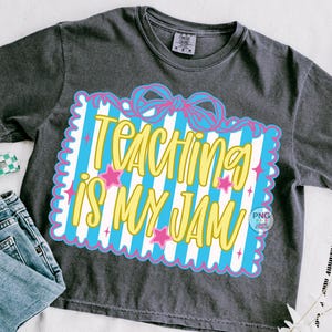 以下が含まれることがあります： 「TEACHING IS MY JAM」の黄色い文字が、青と白のストライプの背景に描かれた濃いグレーのTシャツ。ピンクの星とリボンがデザインされています。Tシャツは青いジーンズと組み合わせています。