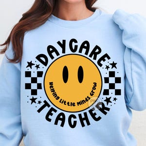 Peut inclure: Sweat-shirt bleu clair avec un motif graphique noir et jaune. Le graphique repr&eacute;sente un visage souriant avec le texte "Daycare Teacher" et "Helping Little Minds Grow" autour du visage.