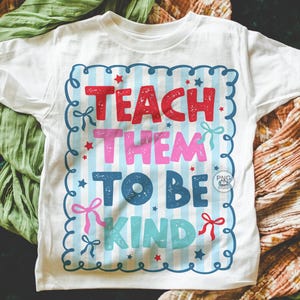 以下が含まれることがあります： 赤、ピンク、青、ターコイズ色の「TEACH THEM TO BE KIND」という文字がプリントされた白いTシャツ。テキストは、リボンと星が付いた青と白のストライプのフレーム内にあります。シャツは柔らかい素材で作られています。