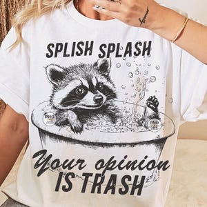 K&ouml;nnte beinhalten: Wei&szlig;es T-Shirt mit einer schwarz-wei&szlig;en Illustration eines Waschb&auml;ren in einer Badewanne mit Blasen. Der Text "Splish Splash Your Opinion Is Trash" ist auf das T-Shirt gedruckt.