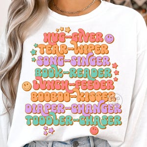 Peut inclure: T-shirt &agrave; manches longues blanche avec un graphique r&eacute;tro color&eacute; qui dit "Hug-Giver, Tear-Wiper, Song-Singer, Book-Reader, Lunch-Feeder, Booboo-Kisser, Diaper-Changer, Toddler-Chaser".