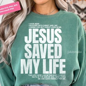 Może przedstawiać: Zielona bluza z napisem "JESUS SAVED MY LIFE" dużymi białymi literami. Bluza ma r&oacute;wnież dodatkowy tekst nad i pod gł&oacute;wnym wyrażeniem. Projekt jest postarzany, co nadaje mu wygląd vintage.