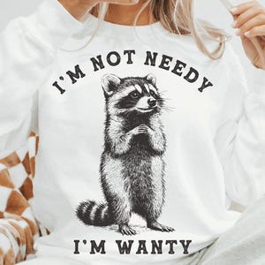 May include: White sweatshirt with a black and white illustration of a raccoon. The text "I'm not needy I'm wanty" is printed on the sweatshirt.
