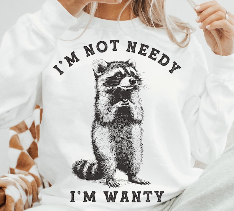 May include: White sweatshirt with a black and white illustration of a raccoon. The text "I'm not needy I'm wanty" is printed on the sweatshirt.